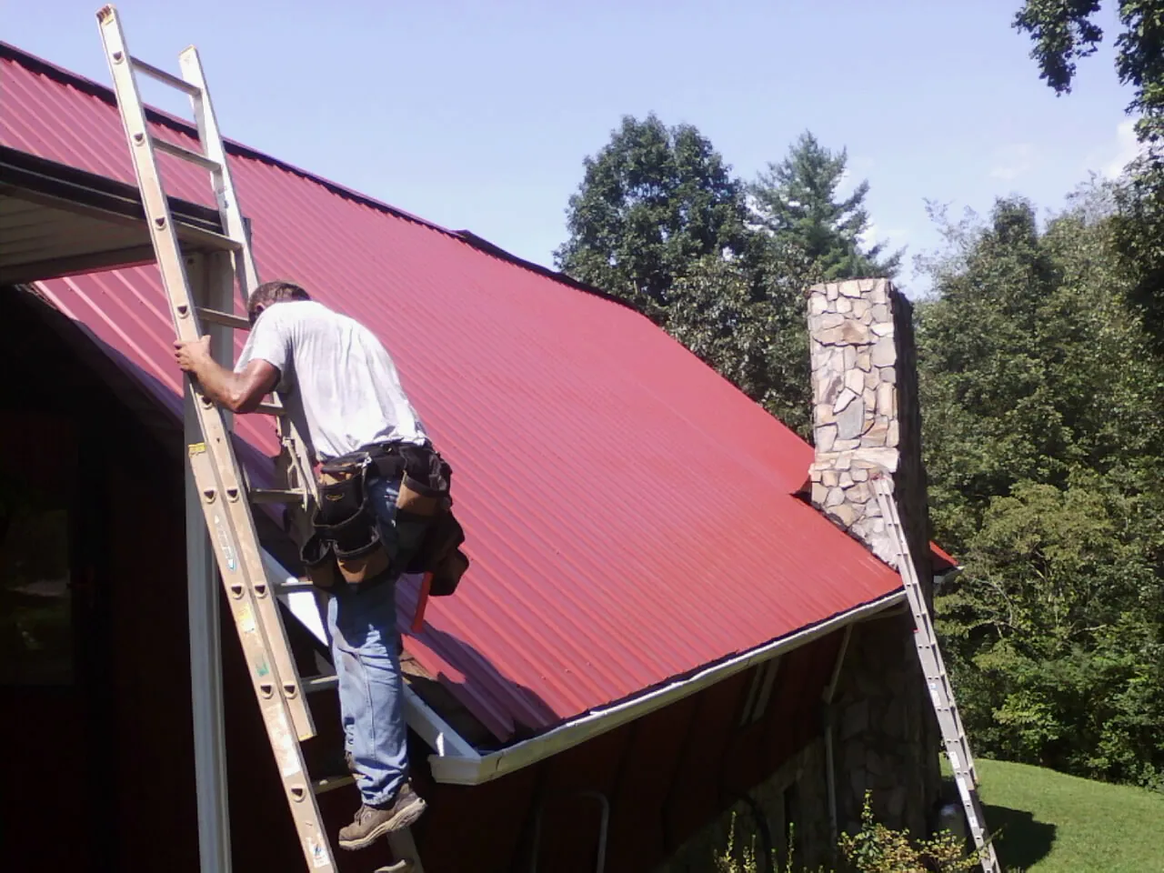 Roofer on a ladder beside a red metal roof and stone chimney — Soffit & Fascia Repair by Roofing Brothers in Endicott
